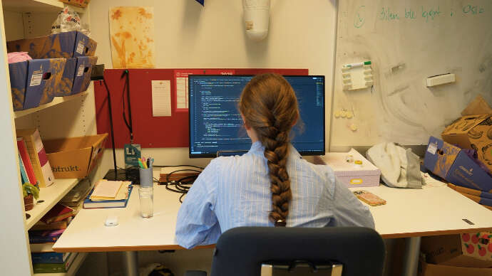 Ung dame med brun, lang flette og lyseblå skjorte sitter ryggen til ser på en pc-skjerm.