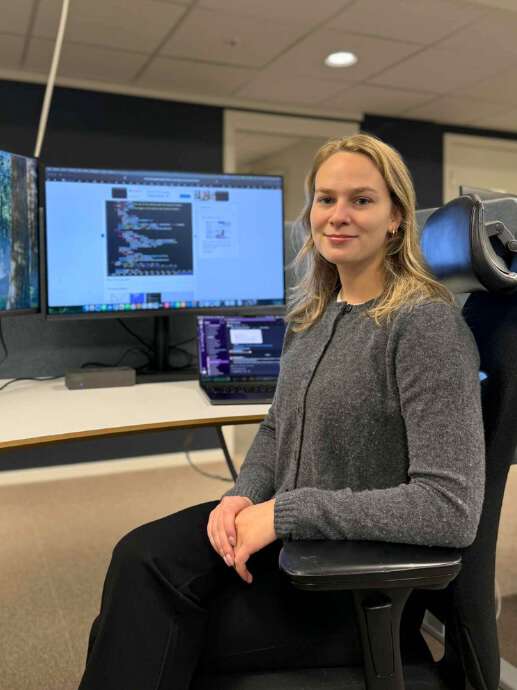 Blond ung dame med grå cardigen sitter foran en pc-skjerm og smiler til kamera.