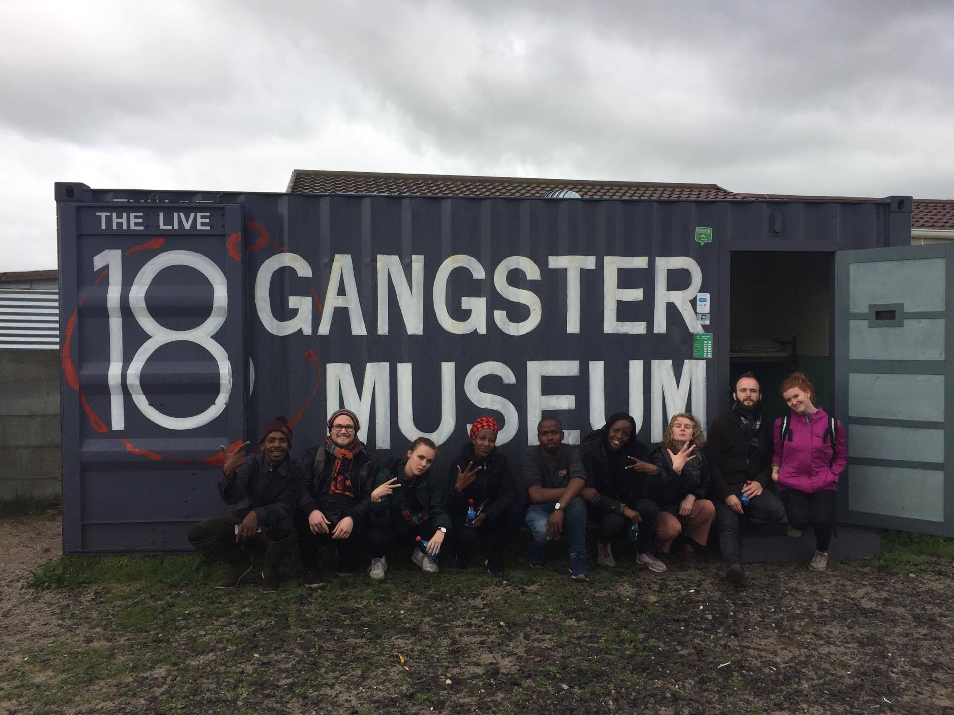 En gruppe mennesker som sitter utenfor en konteiner som det står 18 Gangster Museum på