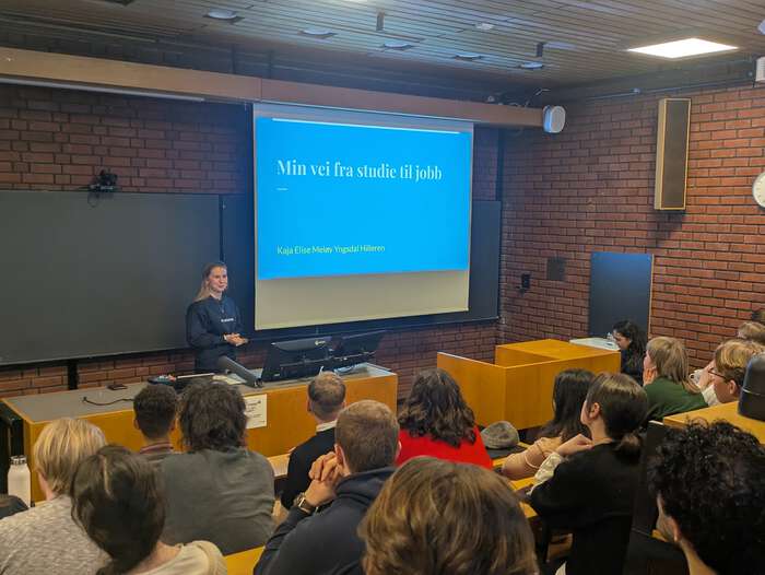 Ung kvinne i forelesningsrom på UiO. Ser presentasjonen "Min vei fra studier til jobb" og bakhoder på de som lytter.