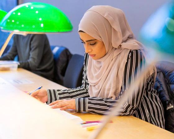 En student sitter ved en pult i Universitetsbibiloteket, en grønn lampe i forgrunnen.