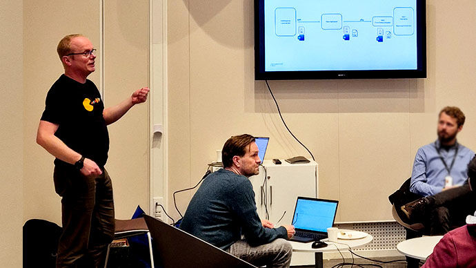 Two men in front of a screen mounted on the wall. The screen displays a flowchart. One man is explaining something, and the other is sitting in front of a laptop.