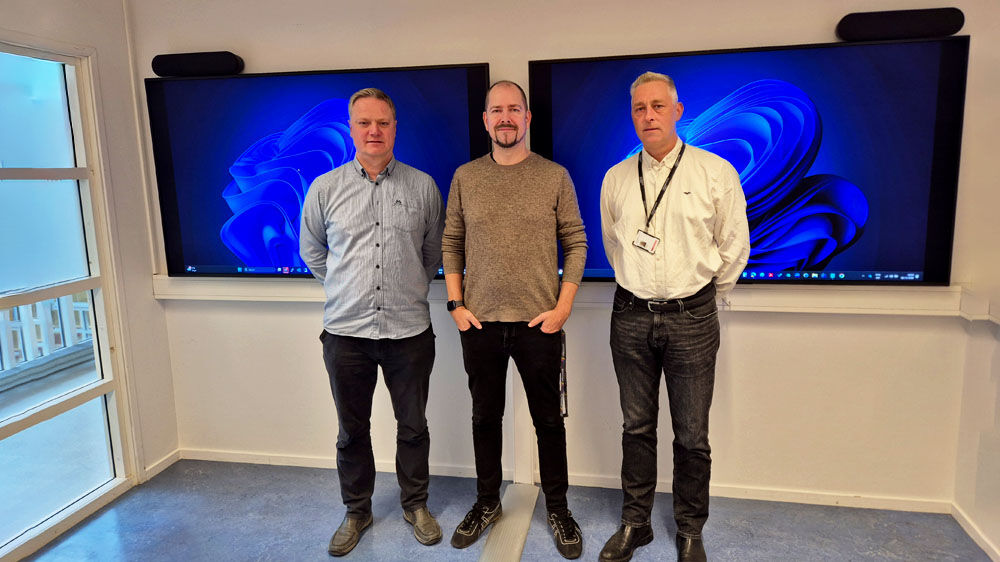 Tre fornøyde menn foran to veggmonterte skjermer som viser et skrivebord med Windows 11