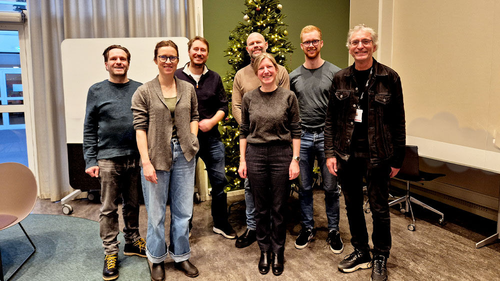 Seven people are standing in front of a board and a Christmas tree and look happy.