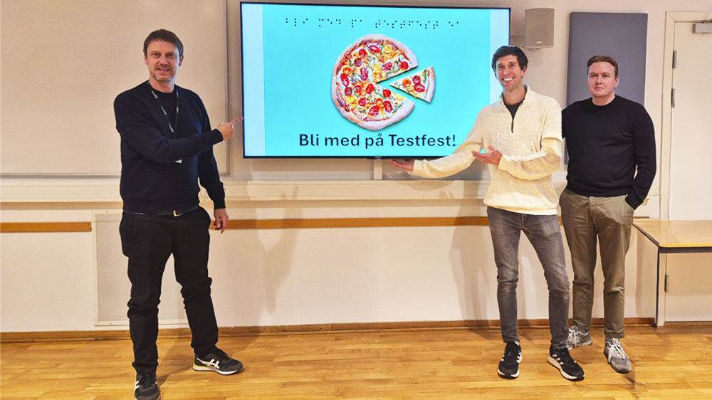 Three men smiling and pointing at a screen with a picture of a pizza and the text Join the test party, both in Latin letters and Braille